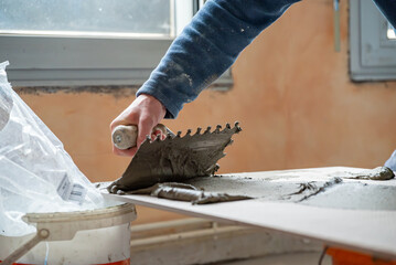 Focus sur la pose de carrelage. Mise en avant d'un homme qui étale la colle sur le carrelage afin de le coller au sol. Gros plan sur l'encollage des carreaux de carrelage.