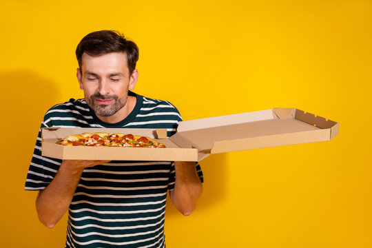 Young man savoring the aroma of a fresh pizza in a yellow background, stylish and casual in a striped t-shirt