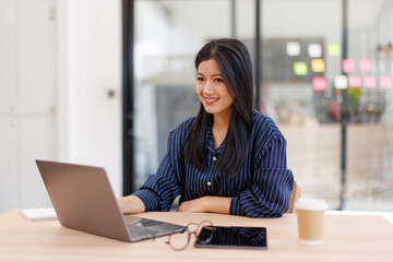  Asian businesswoman sits at desk with laptop online using internet and notepad happily recording...