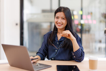  Asian businesswoman sits at desk with laptop online using internet and notepad happily recording...