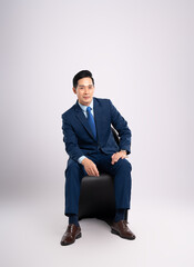 Full body image of Asian male businessman wearing a suit sitting on a chair and posing on a white...