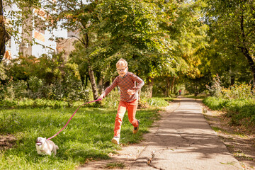 Fototapeta premium Boy in a striped t-shirt and orange pants, wearing glasses with special lenses, running with a small dog on a path in an old park