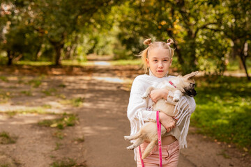 Blonde girl in a white poncho holding a small dog in a warm, sunny, old park