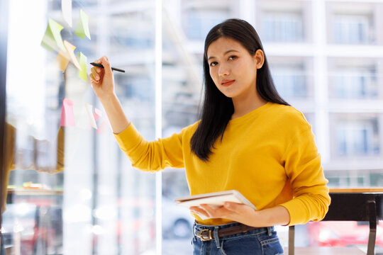 Young asian woman use post it notes to share idea sticky note on glass wall. Asian business people design planning and Brainstorming thinking sticky History notes concept.