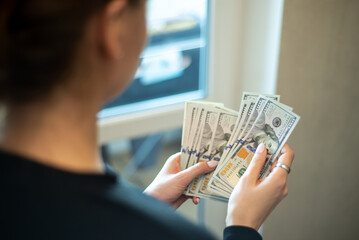 Person counts multiple hundred dollar bills by a window in a modern setting during the daytime