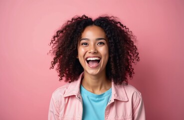 Joyful young woman with curly hair announces news. Casual pink jacket, blue t-shirt. Excited expression, happy face. Beauty portrait on pink background. Smiles, shouts, expressing positive emotion.