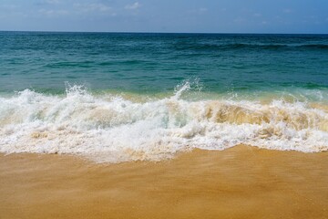 A beautiful sandy beach extends along the coast, with gentle waves lapping the shore. The sun shines in a blue sky, while fluffy white clouds drift above, creating a peaceful atmosphere.