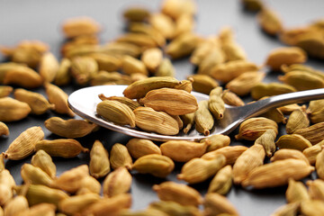 Green cardamom pods in spoon with scattered spices on blurred background. Macro shot. Front view