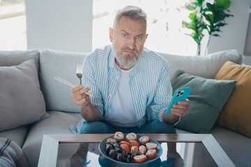 Elderly man dining at home with a plate of sushi, holding a smartphone and chopsticks, expressing a...