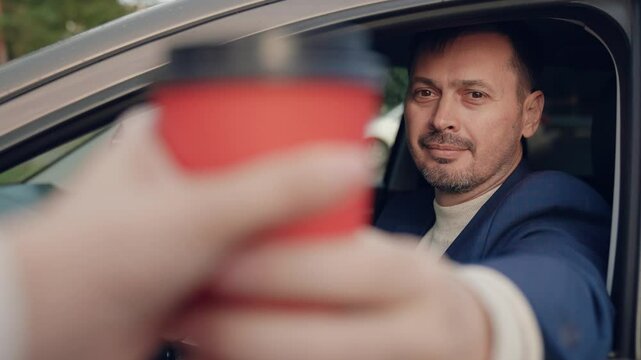Happy driver takes coffee cup from drive-in waitress sitting in car salon. Smiling man orders fresh coffee at drive-in point on city street. Driver drinks hot coffee sitting in modern automobile