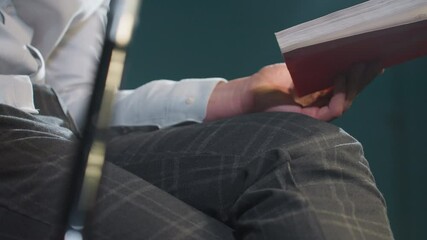 Close-up of faceless person in white shirt and grey trousers holding book and cue stick. Hands rest on lap with fingers gripping cue stick. Soft lighting highlights textures of clothing and objects