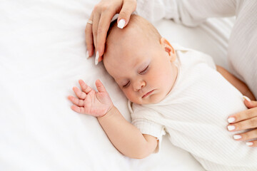 a little baby sleeps sweetly on a white bed, mother's hand gently strokes the baby's head, mother's...