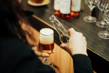 A person pours a golden, frothy beverage into a glass with a gold rim at a bar, creating a warm, inviting atmosphere with blurred bottles and glasses in the background