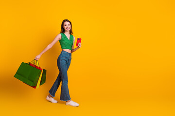 Young cheerful woman holding shopping bags and smartphone, walking confidently on a vibrant yellow...