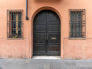 A vintage, arched, wooden door and two windows on an ocher colored wall by the sidewalk. Travel in Italy.
