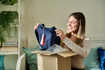 Young woman unpacking cardboard box parcel with online order, looking at new clothes