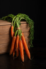 Close-up of a bunch of carrots with green leaves on a wooden crate, black background, vertical with copy space