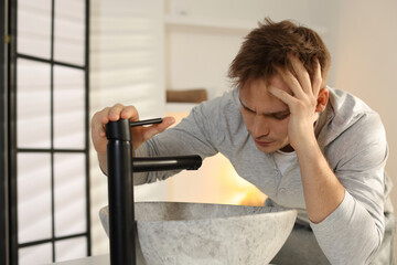 Unhappy young man suffering from hangover near sink in bathroom
