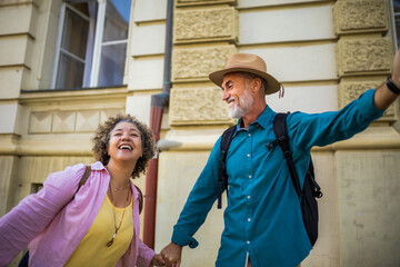 Elderly married couple exploring new city. They are holding hands and wearing hats and backpacks.