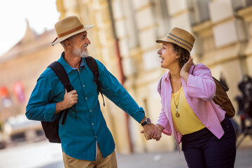 Elderly married couple exploring new city. They are holding hands and wearing hats and backpacks.