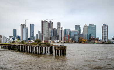 An old pier stands in the Thames River, with the buildings of East London lining the distant bank...