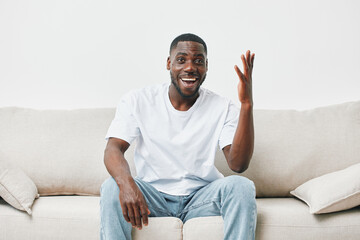 Joyful Black man sitting on a sofa, expressing enthusiasm and engagement, wearing a casual white t...