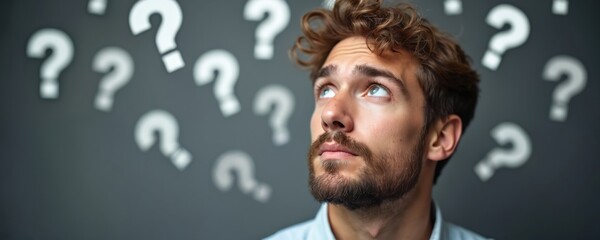 Puzzled caucasian man with beard looks up surrounded by question marks on grey background. He is thinking, solving problems, seeking answers. The guy is looking for ideas, decisions, inspiration.