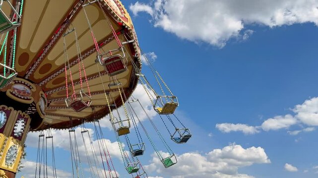 Colorful swing carousel ride at an amusement park under a bright blue sky with white clouds. Empty chairs sway in the wind, creating a sense of excitement and nostalgia