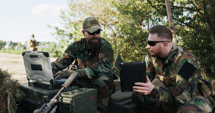 Soldiers in camouflage uniforms, equipped with helmets and backpacks, stand at the training ground, planning a surprise attack, entering data into the system, and holding a tablet while discussing