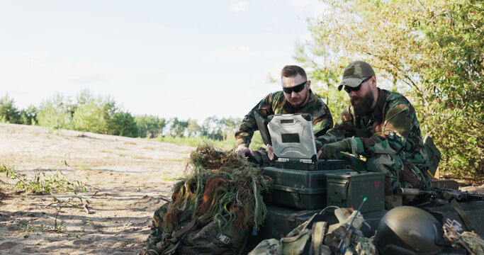 In the field, the army commander prepares reports on his computer to send to the base. A man arrives with a map, and they analyze details as soldiers stand guard in the background.