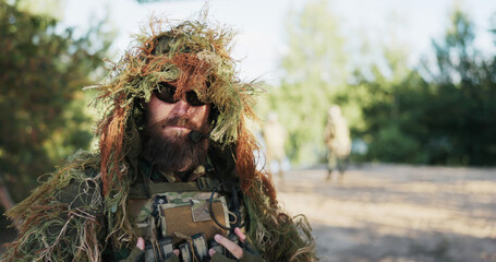 The army commander with a beard wears a moss-covered camouflage cloak and black glasses, preparing...
