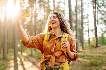 Portrait of a beautiful female hiker with a backpack taking a selfie in a sunny forest. Young woman...