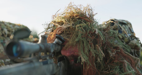 A camouflaged soldier lies in cover, raises his binoculars to observe attackers in the distance,...