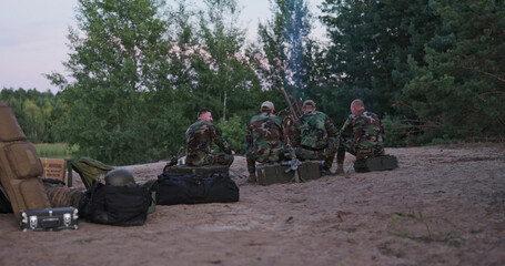 A view from behind shows hunters in camouflage roasting sausages over a bonfire, sharing stories,...