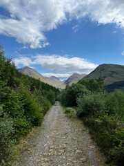 Fototapeta premium A scenic walking path in Glencoe, Scotland, surrounded by lush greenery and towering mountains on a bright spring day.