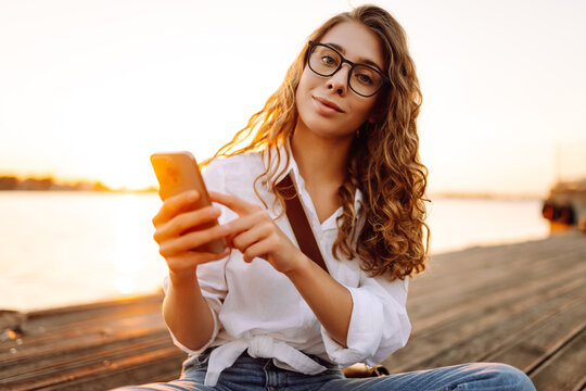 Cute woman walks along the embankment at sunset with a phone in her hands. A beautiful woman with a phone chats outdoors. Blogging, leisure concept. - Powered by Adobe