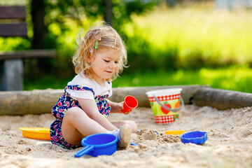 Cute toddler girl playing in sand on outdoor playground. Beautiful baby in red trousers having fun on sunny warm summer day. Child with colorful sand toys. Healthy active baby outdoors plays games
