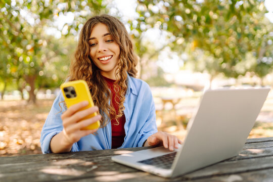 Beautiful woman with phone and laptop sitting at a table in a sunny park. Female freelancer working outdoors. Freelance concept, remote work.