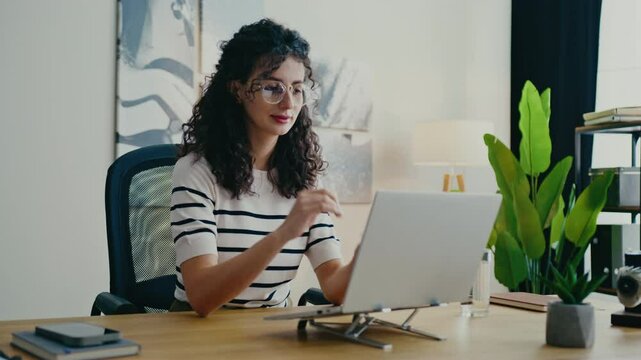 Caucasian girl sitting at office desk and drinking water from glass while looking at laptop screen. Taking short break. Modern workspace with plants and organized shelves. Caring about hydration.