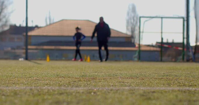 A coach trains a player on a soccer field, with various athletic facilities in the background