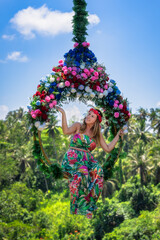 A joyful woman swings on a beautiful floral swing, enveloped by vibrant, lush greenery that vividly captures the essence of an idyllic tropical paradise, Ubud, Bali, Indonesia