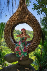 A joyful woman wearing a vibrant floral dress happily poses and shows love sign, in a unique tree nest swing that is beautifully embellished with lush greenery surrounding her, Ubud, Bali, Indonesia