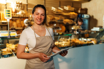 Female baker wearing apron smiling and holding tablet while managing inventory in a bakery with fresh bread and pastries displayed in the background