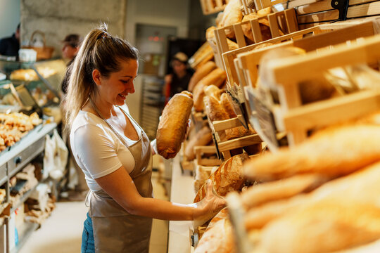 Smiling baker arranging freshly baked loaves of bread on rustic wooden shelves, creating an inviting display that attracts customers in a charming bakery setting - Powered by Adobe