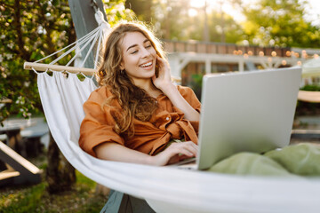Young woman with laptop working lying in a hammock among trees in the fresh air. Female freelancer...