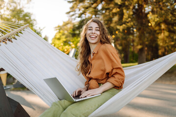 Young woman with laptop working lying in a hammock among trees in the fresh air. Female freelancer...