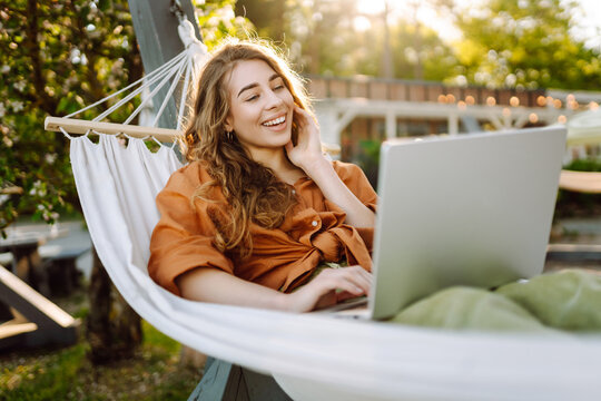 Young woman with laptop working lying in a hammock among trees in the fresh air. Female freelancer spending time in the park at sunset with laptop. Freelance, freedom concept. Active lifestyle.