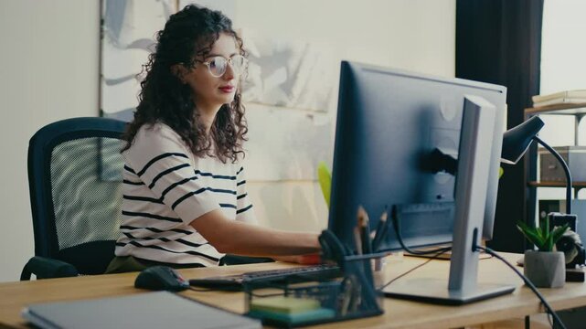Pretty Caucasian girl with curly hair and glasses sitting at desk and applying lipstick while looking at computer screen. Holding cap in one hand and adjusting makeup in bright modern office.