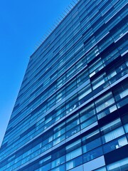 Obraz premium A Modern Glass Building featuring a Reflective Facade, Standing Tall Against a Bright Blue Sky. Modern office building with a sleek glass facade elegantly stands out against a breathtaking blue sky