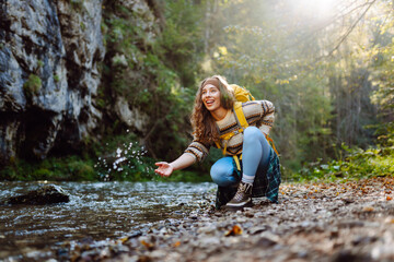 Naklejka premium Young woman with a hiking yellow backpack hiking in the mountains near a mountain river. Happy female traveler enjoying sunny weather outdoors. Travel concept, hiking. Active lifestyle.